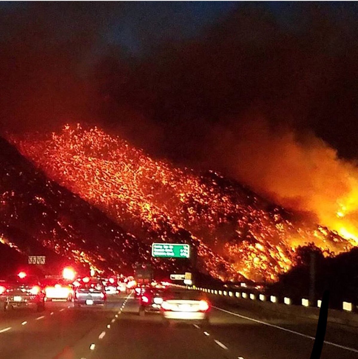 The image shows a number of cars on a freeway, slightly out of focus. It's dark, and the cars all have their lights on. In the background, the world is on fire. The hillside nearest the camera on the left of the image seems to be smouldering, more smoke and coal than fire. Beyond that, you can see a brighter orange, leading to yellow flames on the righthand side of the picture, illuminating the smoke that fills the sky. The cars are all driving straight towards the inferno. It's as if they're commuting into a fiery underworld. 