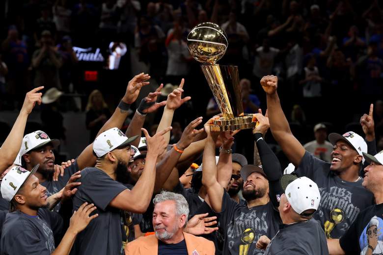New York Knicks players and coaches celebrate on the court after winning the 2026 NBA championship, raising the Larry O’Brien Trophy together while wearing championship hats and gray title shirts following their Game 5 victory over the San Antonio Spurs.