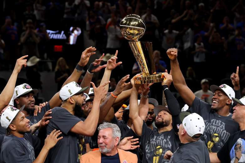 New York Knicks players and coaches celebrate on the court after winning the 2026 NBA championship, raising the Larry O’Brien Trophy together while wearing championship hats and gray title shirts following their Game 5 victory over the San Antonio Spurs.